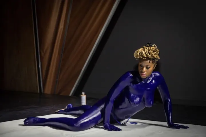Performance of the Women In Culture program: an artist with her body painted blue, on the floor, in a dark exhibition space.